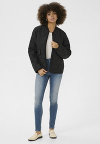 Zwarte gewatteerde bomberjack met een gestructureerd patroon, ritssluiting, zijzakken, gedragen over een licht topje en gecombineerd met blauwe skinny jeans en instappers.