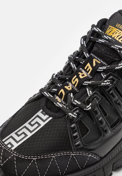 Black athletic shoe with textured mesh upper, rubber accents, and white patterned detailing. Features reflective laces and gold branding.