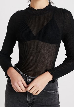 Persoon draagt een doorschijnende zwarte top met lange mouwen over een zwarte bh, gecombineerd met hoog getailleerde vervaagde zwarte jeans. Handen rusten dicht bij de taille.