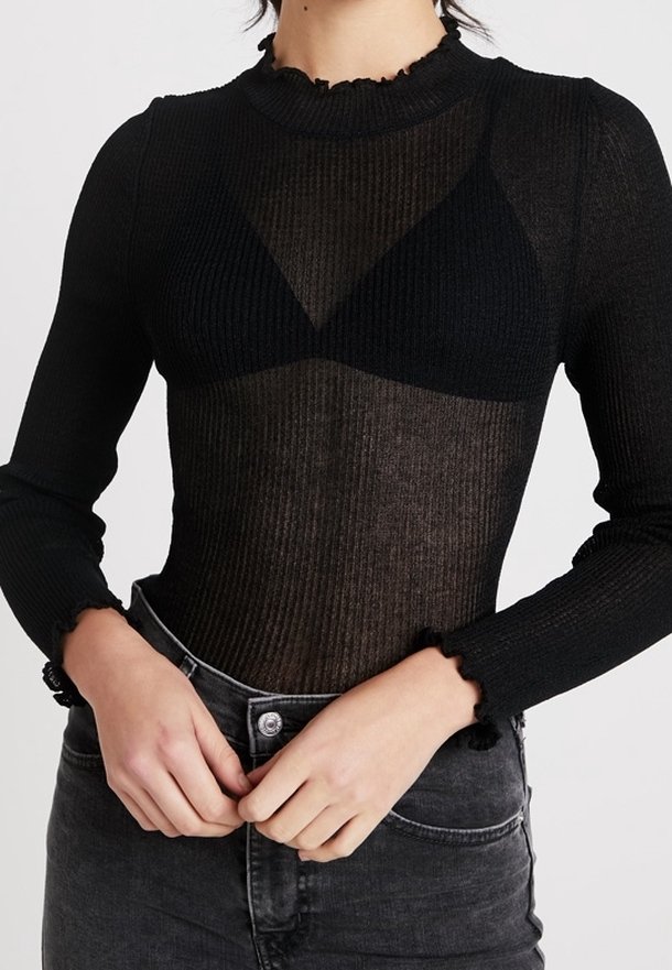 Personne portant un haut noir transparent à manches longues par-dessus un soutien-gorge noir, associé à un jean noir délavé taille haute. Mains reposant près de la taille.