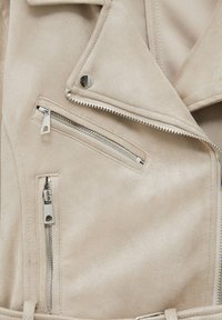 Gros plan sur une veste en daim beige montrant les détails des fermetures éclair argentées, des poches verticales et diagonales, ainsi qu'un bouton-pression sur le col.