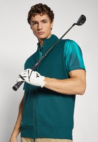 Young man wearing teal golf vest and glove holds golf club resting on shoulder, looking at camera against plain background.