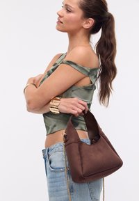 Hnědá měkká suede kabelka se strukturovaným tvarem, krátkým uchem a řetízkovým popruhem. Modelka má na sobě zelený top s designem odhalených ramen.