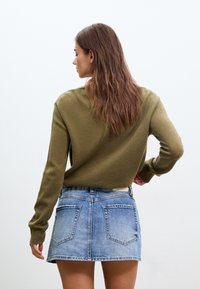 Pull vert olive à manches longues associé à une mini-jupe en denim bleu clair avec deux poches arrière et un ourlet effiloché.
