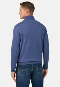Pull en tricot bleu avec un col montant, des poignets et une taille côtelés. Porté avec un jean en denim foncé, affichant une texture lisse et une coupe décontractée.