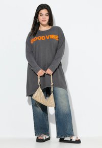 Camisa de manga larga gris con "GOOD VIBE" en naranja, combinada con jeans de mezclilla azul de pierna ancha y sandalias negras. Sosteniendo un bolso metálico pequeño.