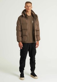 Chaqueta de plumas marrón con capucha, con cremallera frontal, puños elásticos y bolsillos, combinada con una camisa marrón y pantalones cargo negros.