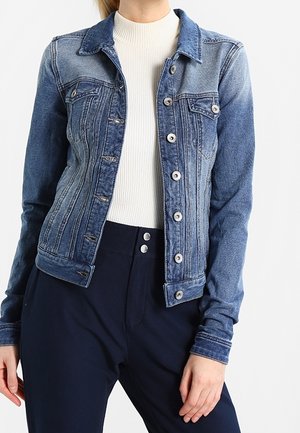 Denimjacka i gradientblått med två bröstfickor, silverfärgad knapprustning och vikta manschetter, kombinerad med en vit ribbad topp och mörka byxor.