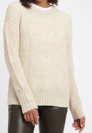 Frau trägt einen cremefarbenen Pullover mit Zopfmuster und schwarze Lederhose, steht vor einem schlichten Hintergrund.