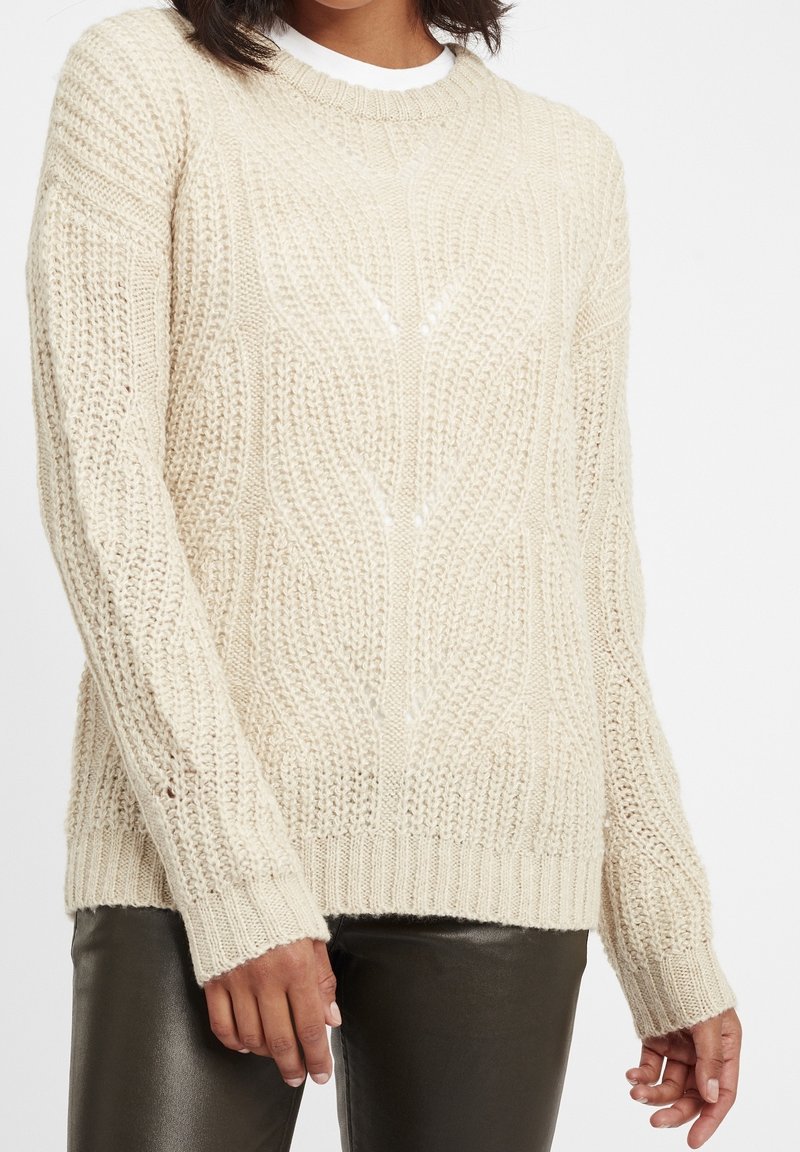 Femme portant un pull en maille torsadée couleur crème et un pantalon en cuir noir, debout devant un fond uni.