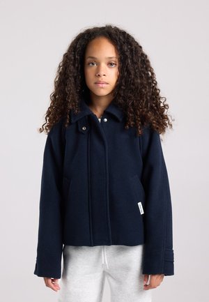 Jeune personne aux cheveux bouclés portant une veste bleu marine et un pantalon de survêtement gris clair, se tenant devant un fond clair uni.