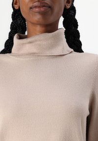 Beige garbó pulóver puha textúrával, bordázott részletekkel a gallérnál és enyhén laza szabással. Zártkörű felvétel, amely a szövetre összpontosít.