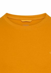 Rundhalsausschnitt, kurzärmeliges T-Shirt in einfarbigem Senfgelb, hergestellt aus weicher Baumwolle mit glatter Textur. Mit kleinem Markenlogo versehen.