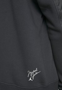 Gros plan sur un tissu noir avec la signature blanche "Michael Jordan" et le logo Jumpman cousus près de l'ourlet sur le bas du vêtement.