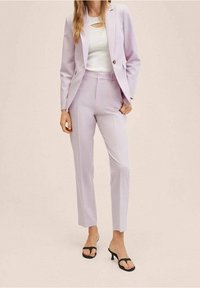 Jasnofioletowy garnitur z dopasowaną marynarką i szytymi na miarę spodniami, w połączeniu z białą bluzką i czarnymi sandałami na obcasie, na neutralnym tle.