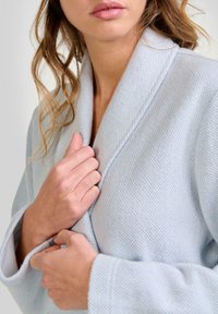 Mujer vistiendo un abrigo texturizado de color azul claro, sosteniendo el cuello con ambas manos cerca del pecho, con el pelo suelto y ondulado a la vista.