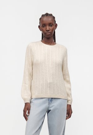 Kvinde med flettet hår iført en cremefarvet striktrøje og lyseblå jeans, stående mod en ensfarvet hvid baggrund.