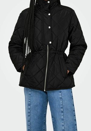 Veste matelassée noire avec fermeture éclair à l'avant et boutons-pression argentés, taille élastique, portée avec un jean bleu, mains dans les poches.