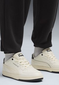 Hvide lædersneakers med sorte accenter, der har perforerede detaljer og sål i sort gummi. Grå ribbede sokker, der stikker op over.
