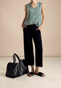 Ärmlös grön och vit mönstrad topp, svarta vida byxor, svart läderväska och svarta slip-in sandaler på en neutral bakgrund.