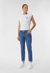 Biała koszulka z krótkim rękawem z grafiką logo, w połączeniu z niebieskimi dżinsami. Dżinsy mają klasyczny krój i podwinięte nogawki, uzupełnione białymi trampkami.