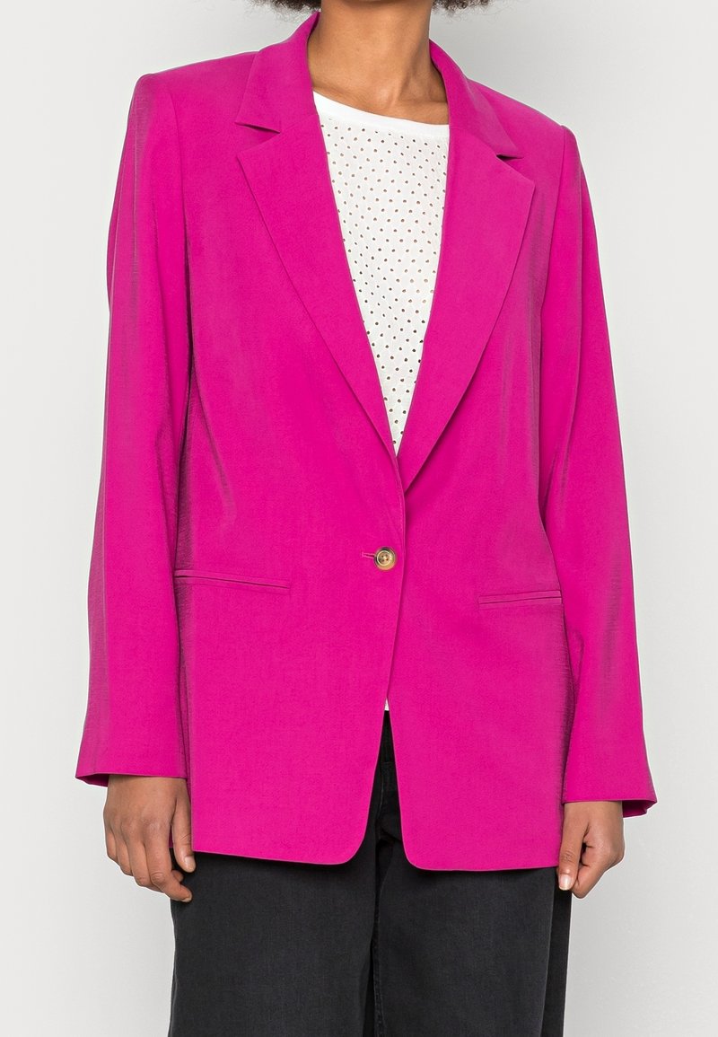 Blazer fucsia con rever a lancia, chiusura con un bottone dorato singolo, due tasche laterali e tessuto liscio; indossato sopra una maglia bianca a pois.
