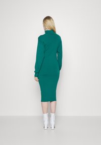 Robe moulante en maille verte avec col haut et manches longues, présentant une texture lisse et une silhouette ajustée, assortie à des bottines blanches.
