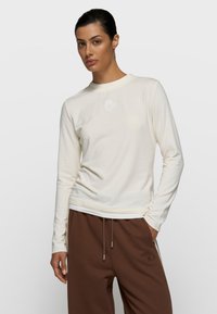 Camisa de manga larga color crema hecha de tela suave, con un pequeño logo blanco en el pecho, combinada con pantalones marrones con cordón.