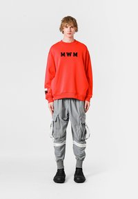 Sudadera roja con "MWM" en negro, combinada con pantalones cargo grises que incluyen cremalleras y correas. Unos zapatos negros completan el look.