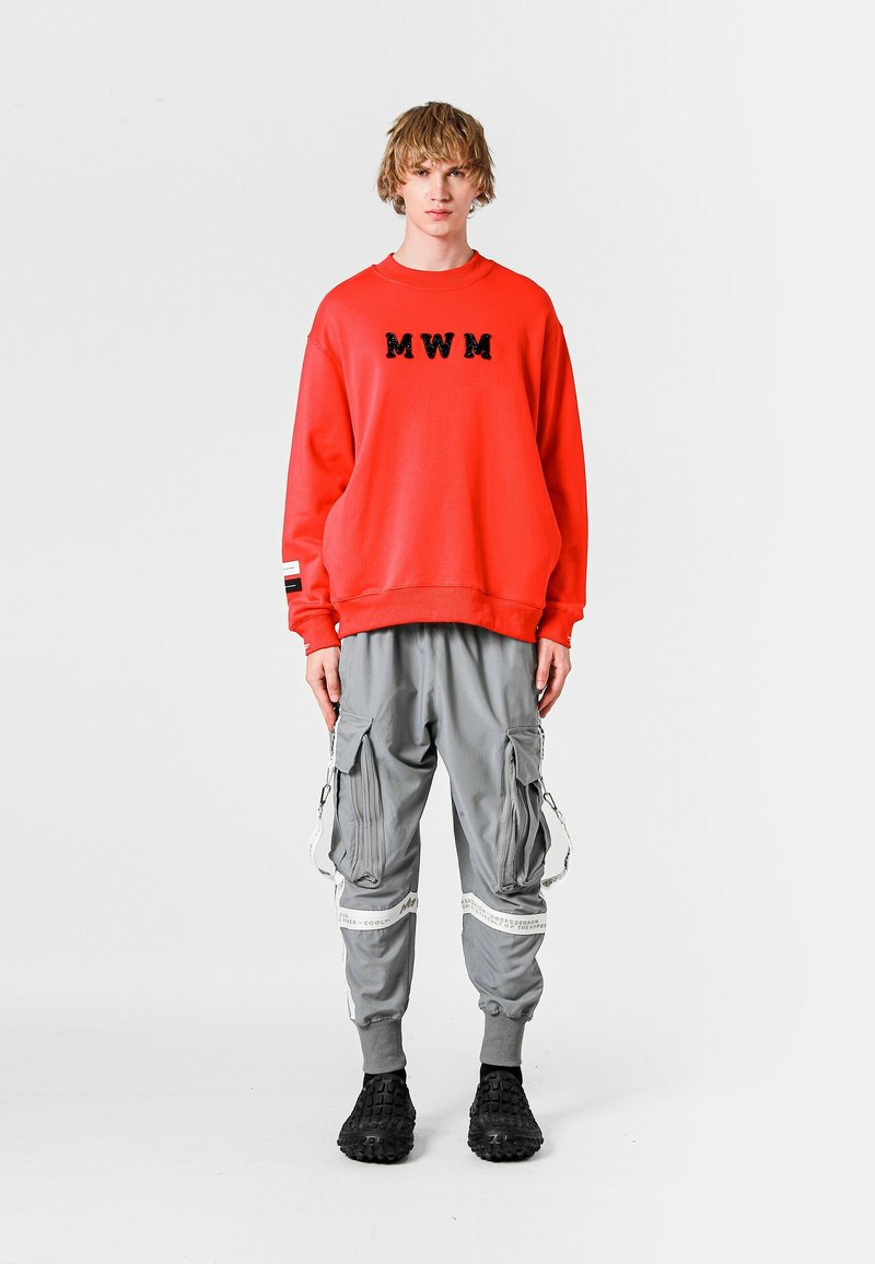 Sudadera roja con "MWM" en negro, combinada con pantalones cargo grises que incluyen cremalleras y correas. Unos zapatos negros completan el look.