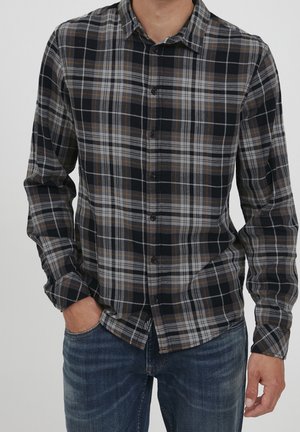 Mann trägt ein schwarz-grau-braunes kariertes Hemd mit Knöpfen und langen Ärmeln sowie blaue Jeans, eine Hand in der Tasche.