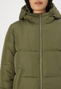 Oliven grønn puffjakke med glidelåslukking, justerbar hette og quiltet design med horisontale seksjoner og sidelommer.