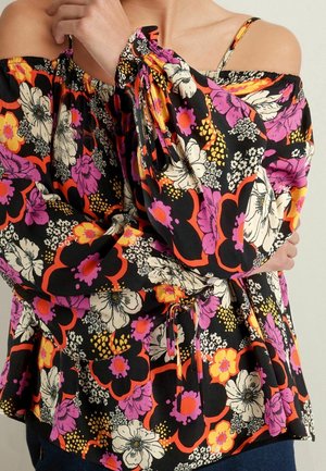 Blouse épaules dénudées avec manches longues, présentant un grand imprimé floral en noir, orange, rose, blanc et jaune, portée par une personne aux bras croisés.