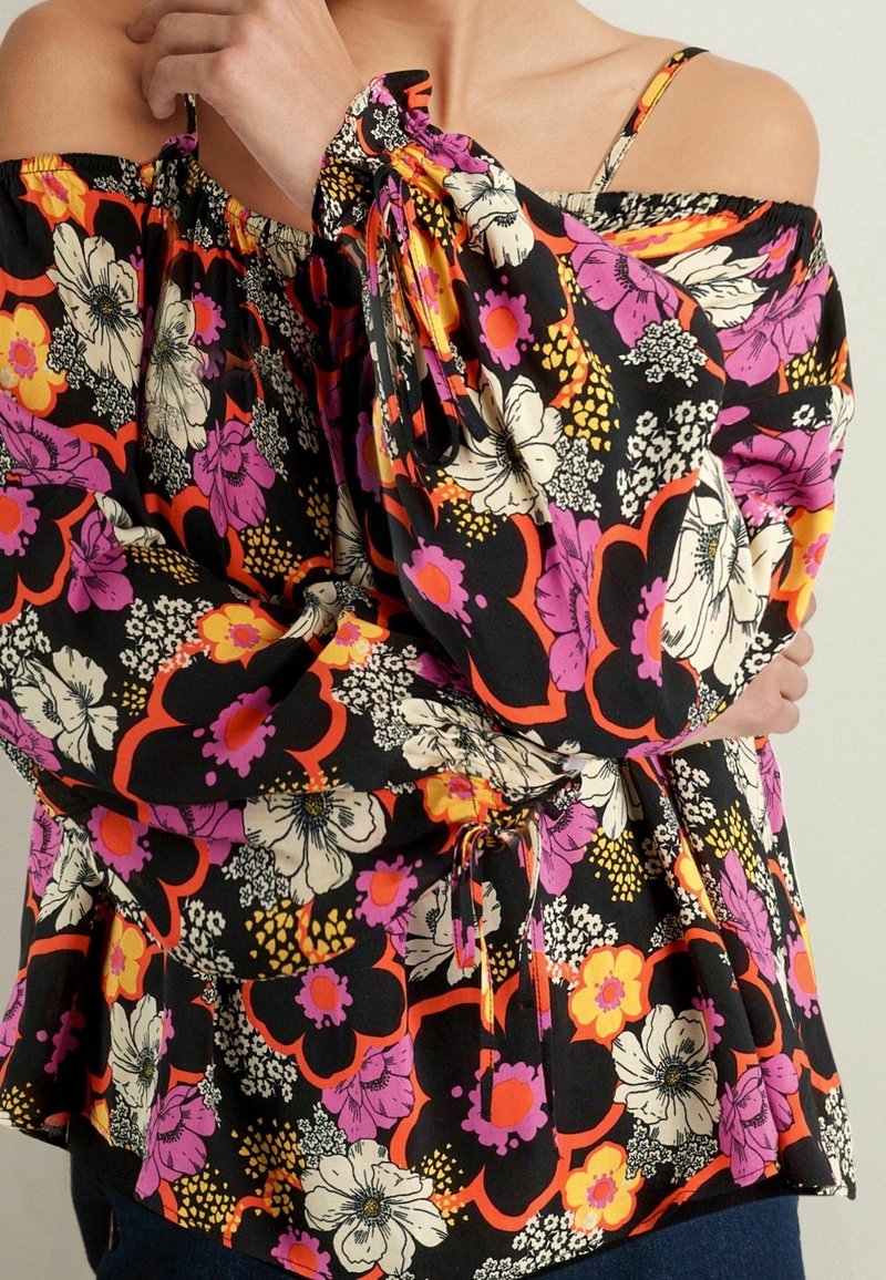 Blouse épaules dénudées avec manches longues, présentant un grand imprimé floral en noir, orange, rose, blanc et jaune, portée par une personne aux bras croisés.