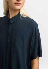 Marineblå kortærmet skjorte med krave, lavet af et silkeagtigt stof. Sømløst design med en let glans og let nedsatte skuldre.