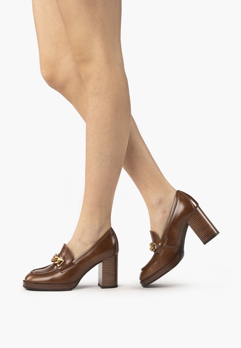 Jambes portant des mocassins en cuir marron à talon bloc avec détail chaîne dorée sur fond blanc, un pied légèrement levé.