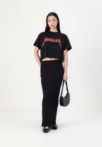 Černé zkrácené tričko s červeným logem "Metallica", spárované s černou, elegantní tužkovou sukní a černými botami, držící puntíkovanou kabelku.