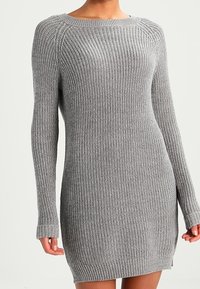 Kvinna som bär en långärmad, ribbad grå stickad klänning med rund halsringning och sidanslits på en enfärgad bakgrund.