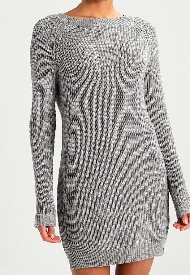 Kvinna som bär en långärmad, ribbad grå stickad klänning med rund halsringning och sidanslits på en enfärgad bakgrund.