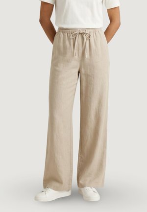 Béžové lněné kalhoty s širokými nohavicemi, elastickým pasem a šňůrkami, nošené s bílými teniskami a bílým krátkým rukávem.