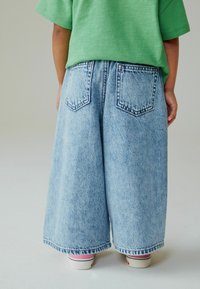 Jean bleu clair à jambes larges en denim avec deux poches arrière, porté avec des chaussures roses et blanches, vu de dos.