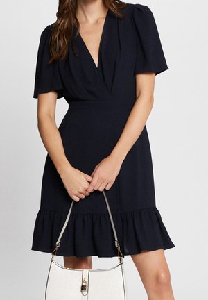 Woman wearing a short black dress with puff sleeves and ruffled hem, holding a white handbag with gold hardware in front.