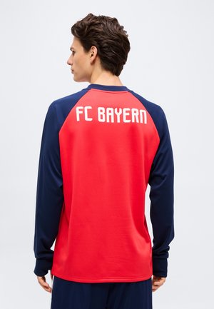 Mand set bagfra iført en rød og marineblå langærmet skjorte med "FC BAYERN" trykt i hvidt på ryggen.