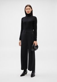 Γυναίκα που στέκεται μπροστά σε ένα απλό φόντο, φοράει μαύρο ζιβάγκο, φαρδιά μαύρη παντελόνα, μαύρα μποτάκια αστραγάλου και κρατά μια μικρή μαύρη τσάντα.