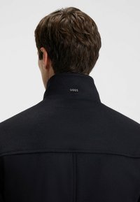 Mann mit kurzen braunen Haaren, der einen schwarzen Mantel mit aufgestelltem Kragen trägt, auf dem hinten ein kleines metallisches "BOSS"-Logo zu sehen ist.
