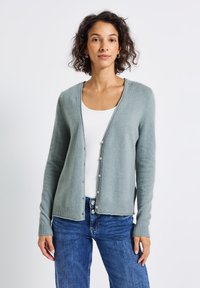 Cardigan vert clair en matériau tricot doux, avec un col en V, fermeture à boutons et ourlet côtelé. Associé à un débardeur blanc et un jean bleu.