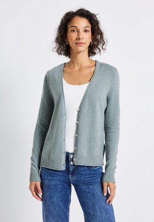 Light green cardigan made of soft knit material, featuring a V-neck, button closure, and ribbed hem. Paired with a white tank and blue jeans.