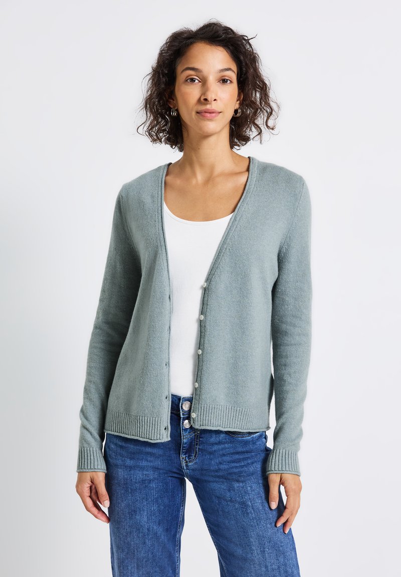 Cardigan vert clair en matériau tricot doux, avec un col en V, fermeture à boutons et ourlet côtelé. Associé à un débardeur blanc et un jean bleu.