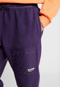Fioletowe dresowe spodnie poliestrowe z fakturowanym wykończeniem i elastycznym paskiem w talii, z połyskującą kieszonką naszytą z tkaniny z czarną metką logo.