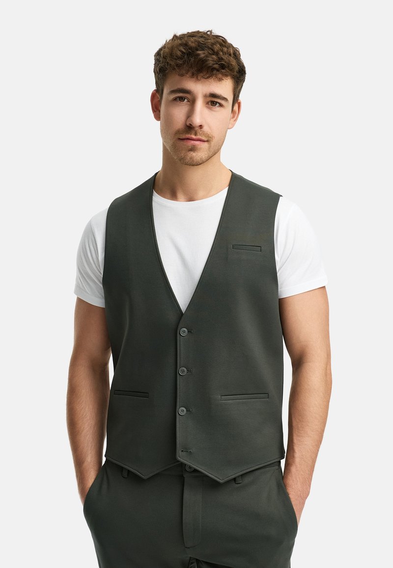 Mørkegrønn vest med en skreddersydd passform, med V-hals design, fem knapper og to frontlommer. Bar over en hvit T-skjorte.